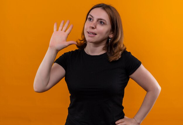 Jonge toevallige vrouw die hallo gebaren en naar linkerkant met hand op taille op geïsoleerde oranje muur kijkt