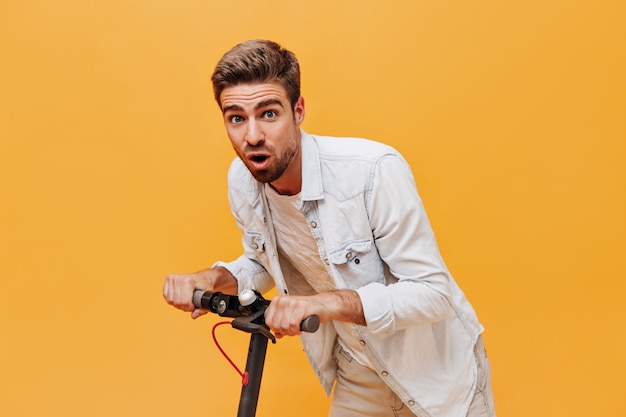 Gratis foto jonge trendy bebaarde man met blauwe ogen in spijkerjasje en geruit t-shirt op zoek naar camera en poseren met scooter op oranje muur