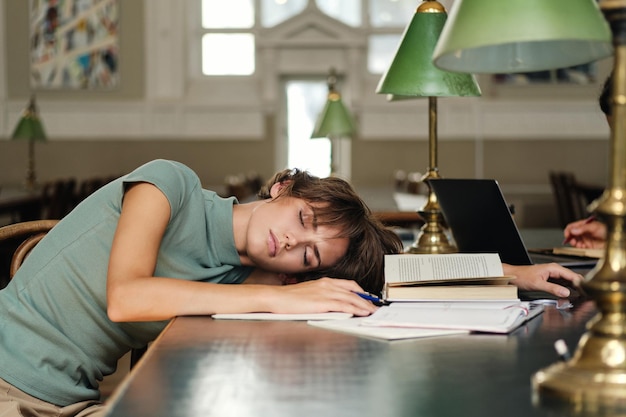 Jonge trieste, vermoeide vrouwelijke student die op een bureau slaapt met boeken in de buurt tijdens studie in de bibliotheek van de universiteit
