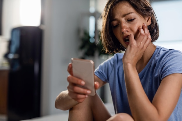 Jonge trieste vrouw die aan iets denkt terwijl ze alleen in de slaapkamer zit en naar haar mobiele telefoon kijkt