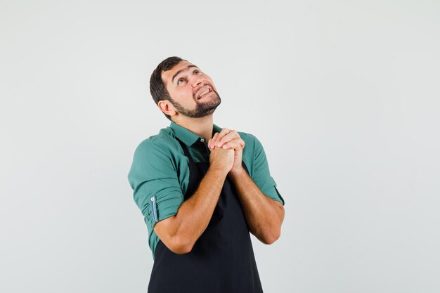 Jonge tuinman die biddend gebaar in t-shirt, schort toont en hoopvol kijkt. vooraanzicht.