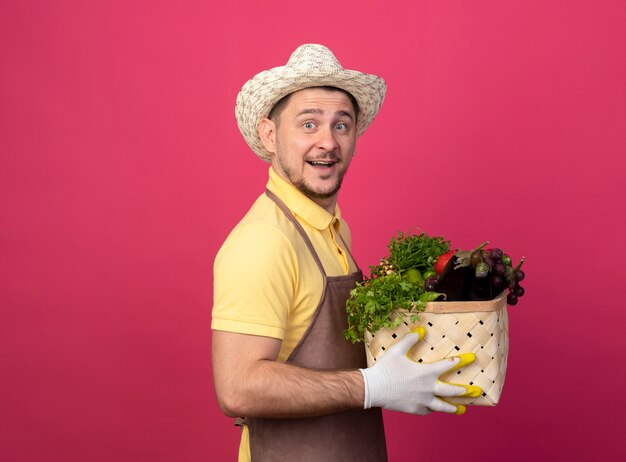 Jonge tuinman die jumpsuit en hoed in werkhandschoenen draagt die een krat vol groenten houdt die aan de voorkant glimlachend met een blij gezicht over roze muur kijkt