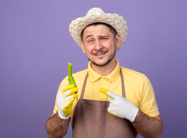 Jonge tuinman die jumpsuit en hoed in werkhandschoenen draagt die gebroken groene chilipeper houdt die met wijsvinger ernaar richt en glimlachend zelfverzekerd over paarse muur staat