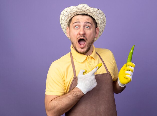 Jonge tuinman die jumpsuit en hoed in werkhandschoenen draagt die groene chilipeper met wijsvinger ernaar houdt, bing verbaasd en verrast staande over paarse muur