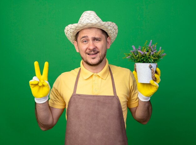 Jonge tuinman die jumpsuit en hoed in werkhandschoenen draagt die ingemaakte installatie over groene muur houden