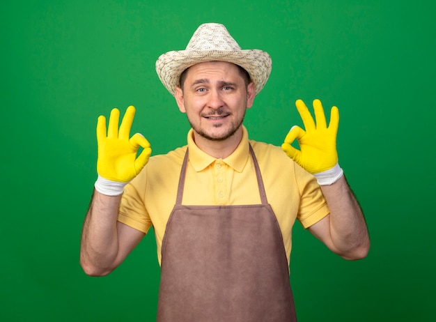 Jonge tuinman die jumpsuit en hoed in werkhandschoenen draagt die voorzijde met gelukkig gezicht bekijkt dat ok teken toont dat zich over groene muur bevindt