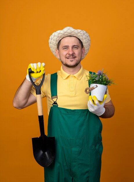 Jonge tuinman in jumpsuit en hoed met schop en potplant kijken naar voorkant lachend met blij gezicht staande over oranje muur