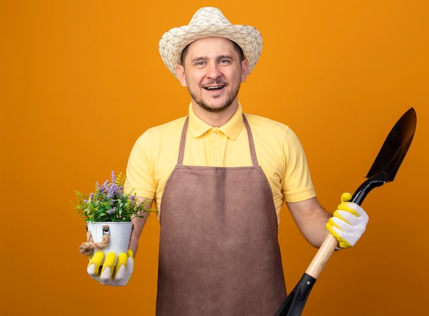 Jonge tuinman in jumpsuit en hoed met schop en potplant kijken naar voorkant lachend met blij gezicht staande over oranje muur Gratis Foto