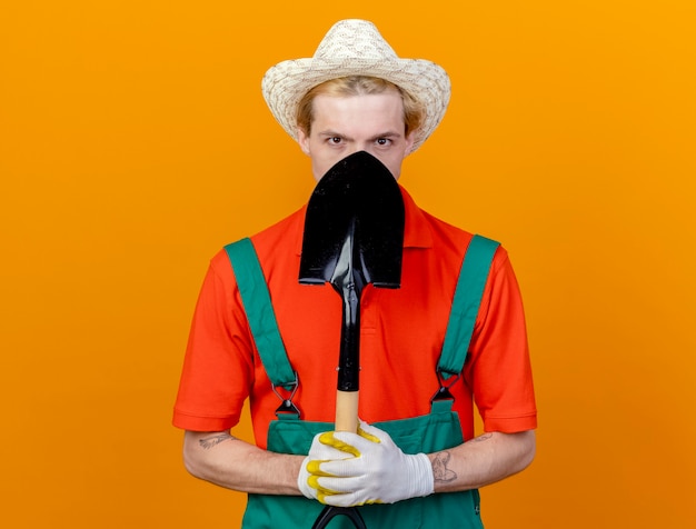 Jonge tuinman man dragen jumpsuit en hoed houden schop kijken camera verbergen zijn gezicht staande over oranje achtergrond