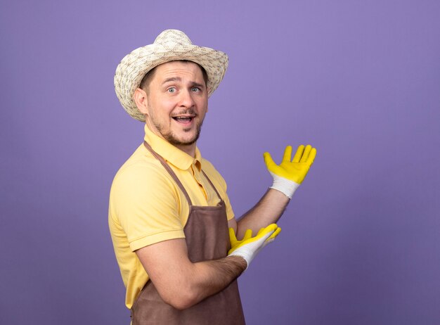 Jonge tuinman man dragen jumpsuit en hoed in werkhandschoenen iets presenteren met armen vrolijk lachend