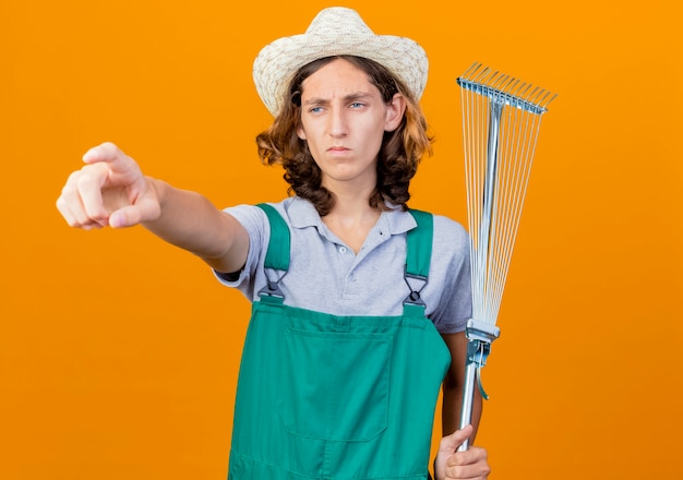 Gratis foto jonge tuinman man dragen jumpsuit en hoed met hark wijzend met wijsvinger