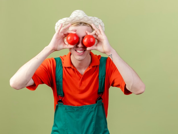 Jonge tuinman man dragen jumpsuit en hoed met verse tomaten bedekken ogen met hen glimlachend staande over lichte achtergrond