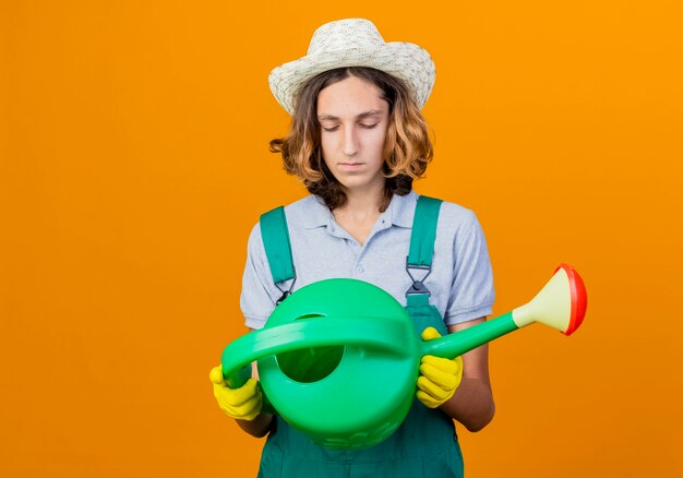 Jonge tuinman man in rubberen handschoenen dragen jumpsuit en hoed met gieter kijken met ernstig gezicht staande over oranje achtergrond