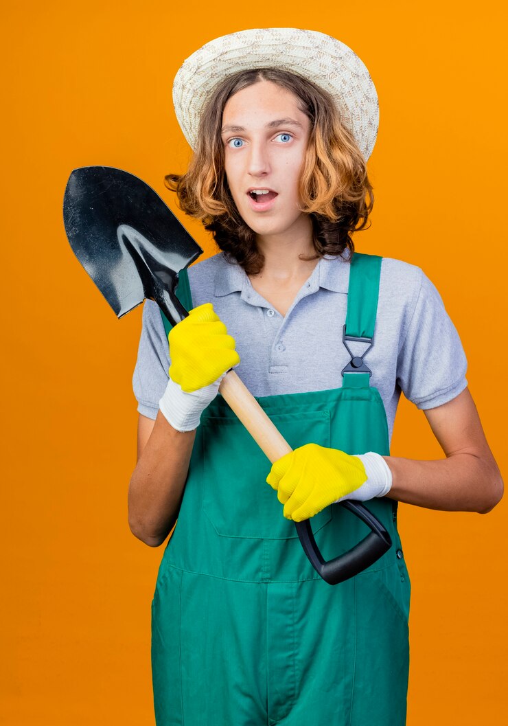 Gratis Foto jonge tuinman man in rubberen handschoenen dragen jumpsuit en hoed met schop glimlachen