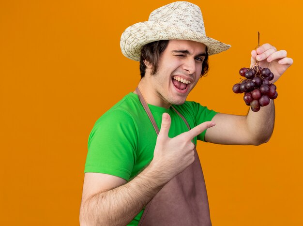 Jonge tuinman man in schort en hoed tonen bos druiven wijzend met index figner ernaar glimlachend en knipogend staande over oranje muur