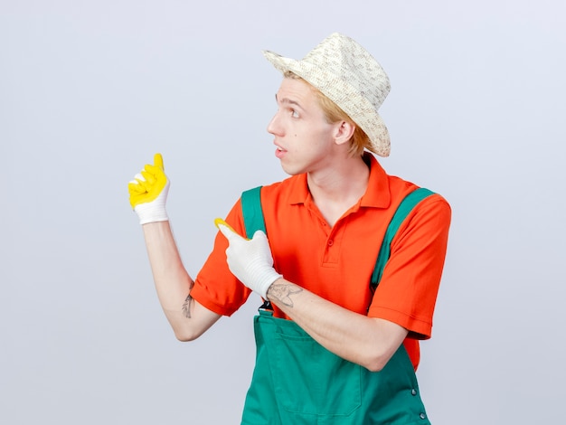 Jonge tuinman man met jumpsuit en hoed in rubberen handschoenen wijzend met wijsvinger naar de zijkant