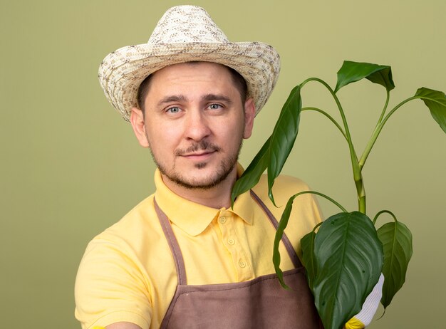 Jonge tuinman man met jumpsuit en hoed in werkhandschoenen bedrijf plant kijken voorkant met glimlach op gezicht staande over lichte muur