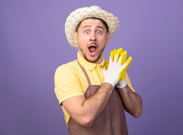 Jonge tuinman man met jumpsuit en hoed in werkhandschoenen hand in hand samen kijken verbaasd en verrast