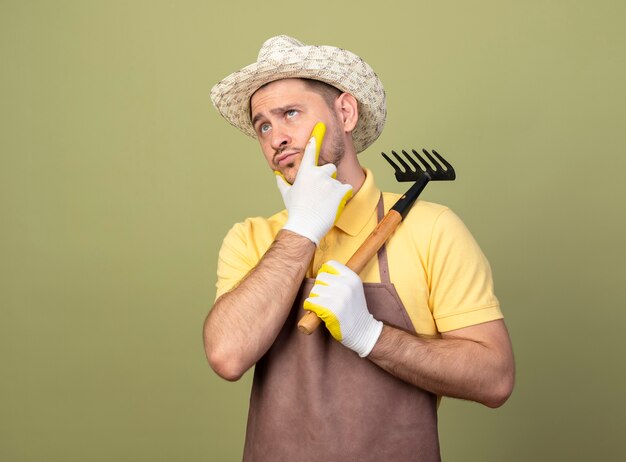 Jonge tuinman man met jumpsuit en hoed in werkhandschoenen met mini hark opzij kijken met hand op kin denken