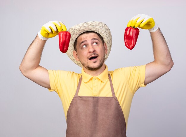 Jonge tuinman man met jumpsuit en hoed in werkhandschoenen met rode paprika glimlachend vrolijk