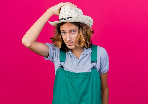 Jonge tuinman man met jumpsuit en hoed op zoek verward zijn hoed aan te raken