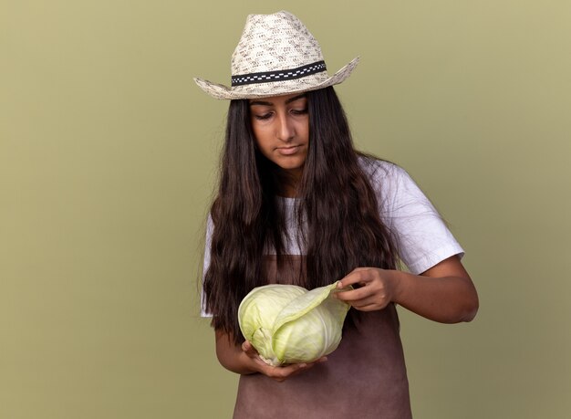 Jonge tuinman meisje in schort en zomer hoed houden kool kijken geïntrigeerd staande over groene muur