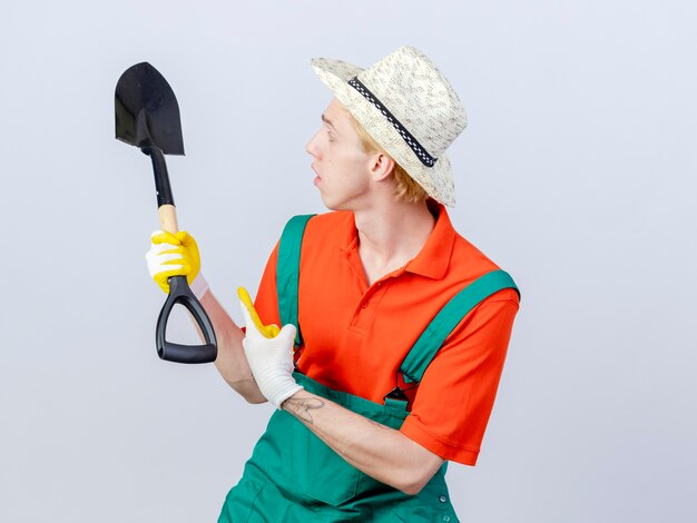 Jonge tuinman met jumpsuit en hoed in rubberen handschoenen met schop die met wijsvinger erop wijst en er verward uitziet