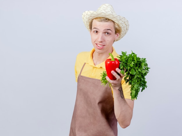 Jonge tuinman met jumpsuit en hoed met rode paprika en verse kruiden met een glimlach op het gezicht