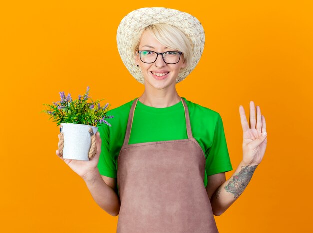 Jonge tuinman vrouw met kort haar in schort en hoed bedrijf potplant kijken camera glimlachend met blij gezicht weergegeven: nummer vier staande over oranje achtergrond
