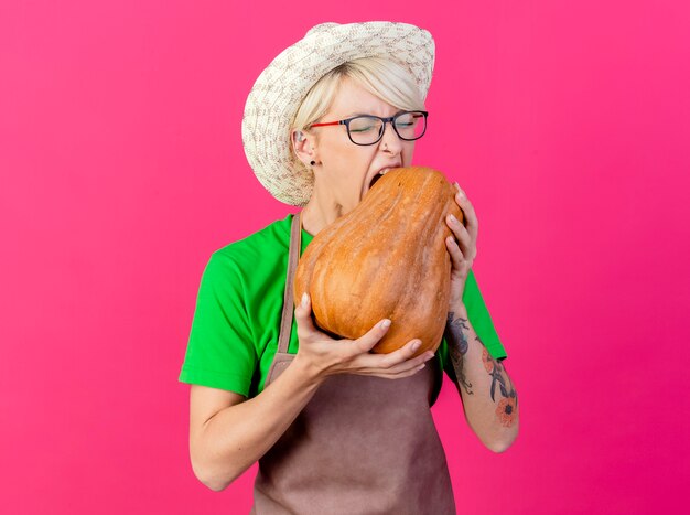 Jonge tuinman vrouw met kort haar in schort en hoed met pompoen proberen te bijten