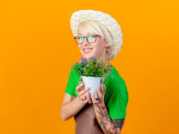 Jonge tuinman vrouw met kort haar in schort en hoed met potplant kijken camera lachend met blij gezicht staande over oranje achtergrond