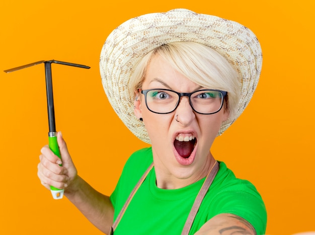 Gratis foto jonge tuinman vrouw met kort haar in schort en hoed swingende mattock schreeuwen met boos gezicht gefrustreerd staande over oranje achtergrond