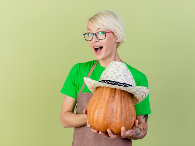 Gratis foto jonge tuinman vrouw met kort haar in schort met pompoen met hoed erop camera kijken met glimlach op gezicht staande over lichte achtergrond