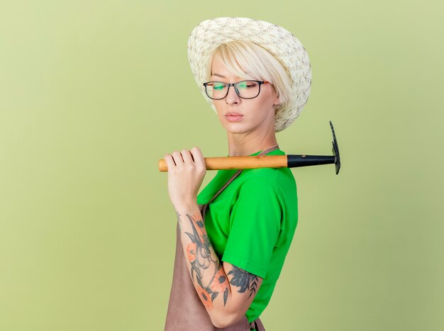 Jonge tuinmanvrouw met kort haar in schort en hoed die minihark op schouder houden die zich zelfverzekerd over lichte achtergrond bevinden