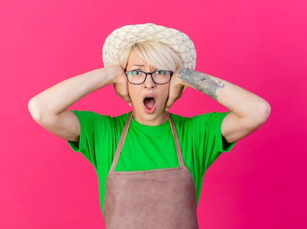 Gratis foto jonge tuinmanvrouw met kort haar in schort en hoed die oren behandelen met handen