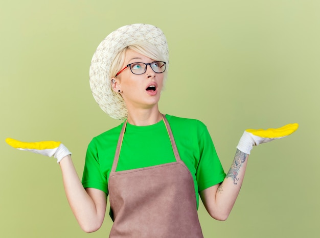 Gratis foto jonge tuinmanvrouw met kort haar in schort en hoed die rubberhandschoenen dragen die zich over lichte achtergrond bevinden