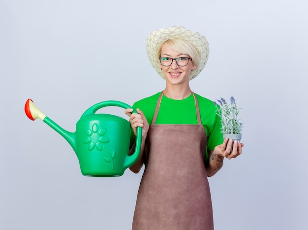 Gratis foto jonge tuinmanvrouw met kort haar in schort en hoed met gieter en potplant glimlachend met een blij gezicht