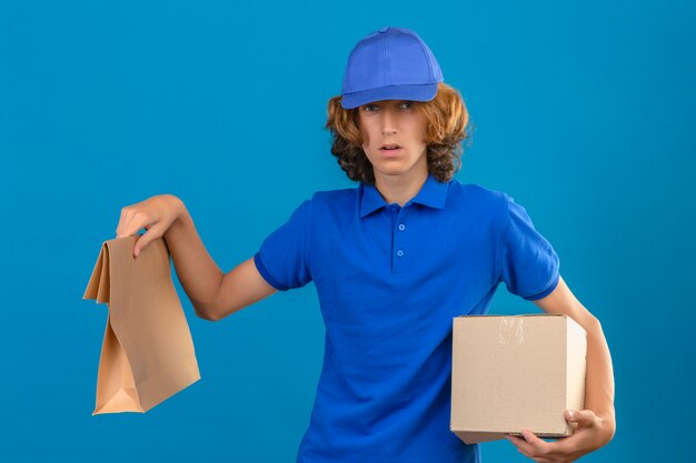 Jonge twijfelachtige leveringsmens die blauw poloshirt en GLB draagt die kartondoos en document pakket houden die camera met twijfels bekijken die zich over geïsoleerde blauwe achtergrond bevinden