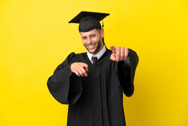 Jonge, universitair afgestudeerde blanke man geïsoleerd op gele achtergrond wijst vinger naar je terwijl hij lacht Premium Foto