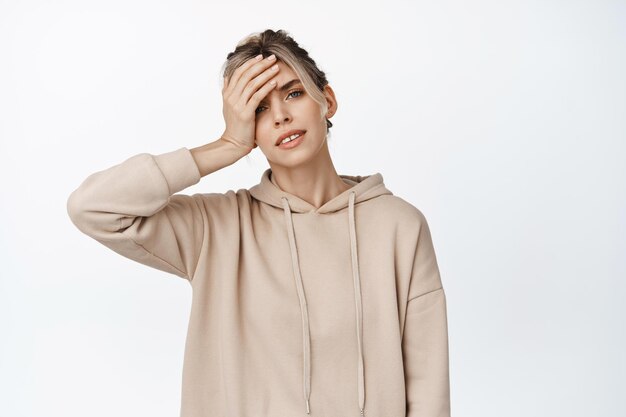 Jonge, vermoeide vrouw die de hand op het hoofd houdt van vermoeidheid met koorts of hoofdpijn in een beige hoodie op een witte achtergrond