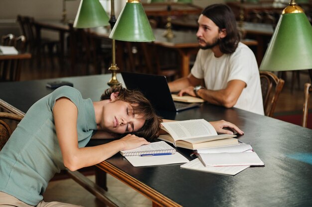 Jonge vermoeide vrouwelijke student slapen op bureau met boeken tijdens studie in bibliotheek van universiteit