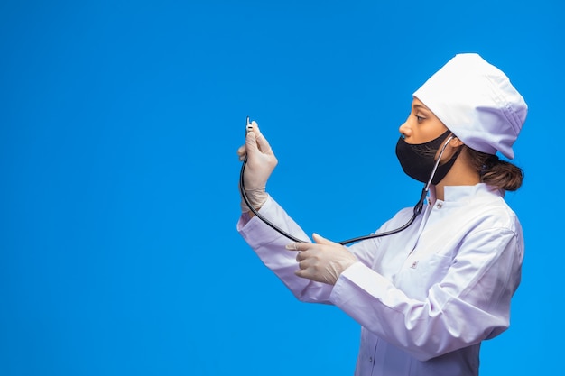 Jonge verpleegster in gezichtsmasker en handhandschoenen controleert de patiënt met een stethoscoop.