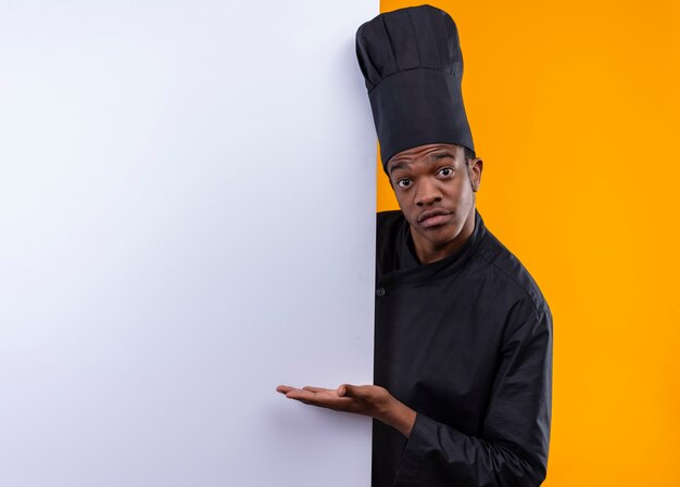 Jonge verrast Afro-Amerikaanse kok in uniform chef staat achter witte muur en wijst naar muur met hand geïsoleerd op oranje muur