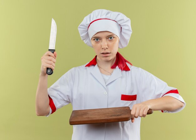 Jonge verrast blonde vrouwelijke chef-kok in uniform chef houdt mes en snijplank geïsoleerd op groene muur