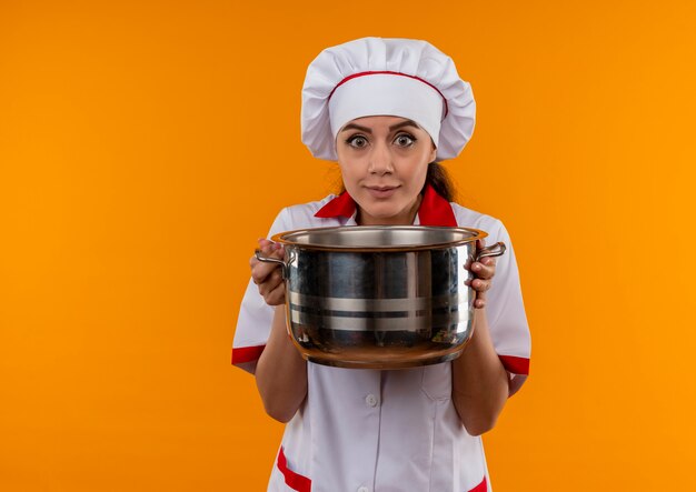 Jonge verrast Kaukasisch kok meisje in uniform chef houdt pot en kijkt naar camera geïsoleerd op oranje muur met kopie ruimte