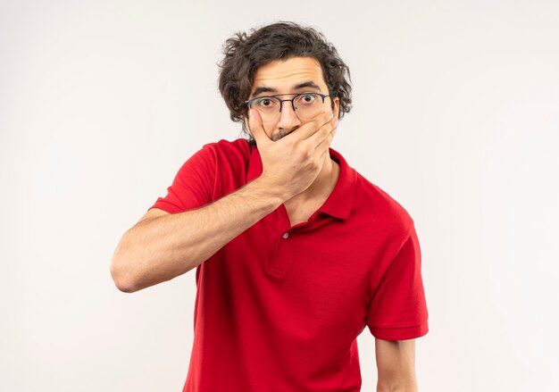 Jonge verrast man in rood shirt met optische bril legt hand op de mond en kijkt geïsoleerd op een witte muur