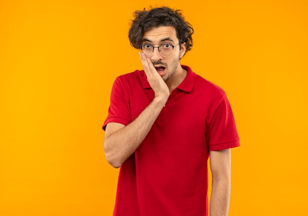 Jonge verrast man in rood shirt met optische bril legt hand op kin en kijkt geïsoleerd op oranje muur