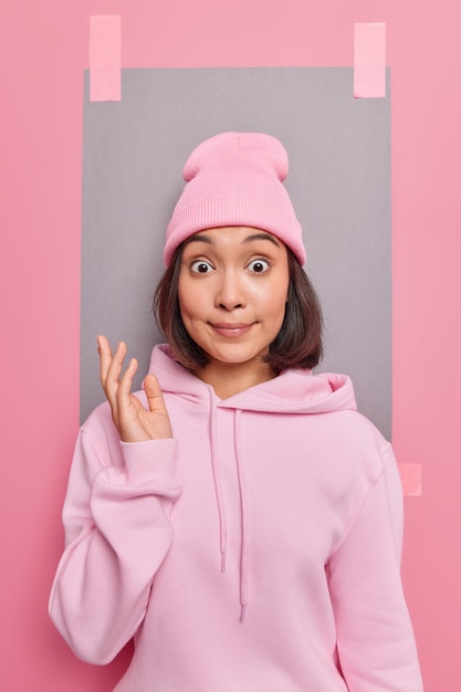 Jonge verraste jonge aziatische vrouw staart afgeluisterde ogen houdt hand omhoog met verontwaardiging draagt casual sweatshirt en hoed poses tegen roze achtergrondgeluid met lege lege ruimte voor uw advertentie