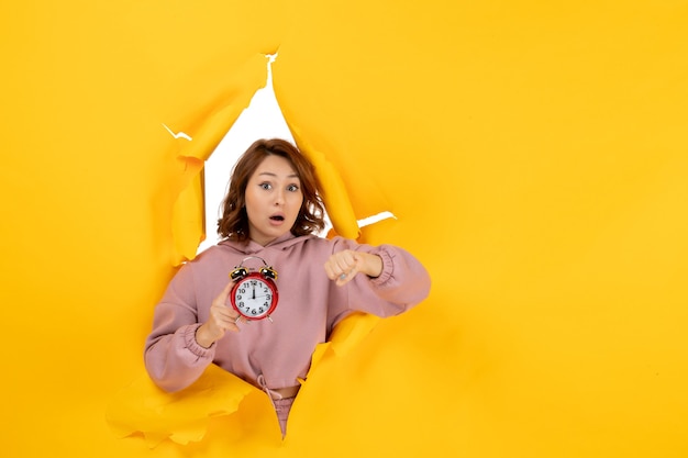Gratis foto jonge verraste mooie dame die klok vasthoudt en haar tijd controleert op gele gescheurde doorbraakachtergrond