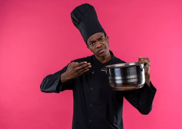 Jonge verwarde Afro-Amerikaanse kok in uniform chef houdt steelpan en punten met hand geïsoleerd op roze muur